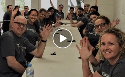 A group of people sit along a long set of tables, all leaning in and waving down the corridor they create. The image has a play button on top showing it is a screen capture from a video.