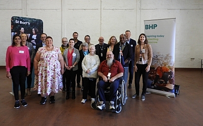 A diverse group of people gather together in a hall in front of two banners, one has St Bart's on it, the other BHP