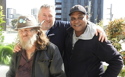 Three men stand together with their arms wrapped around each other. They are outside among city buildings and greenery
