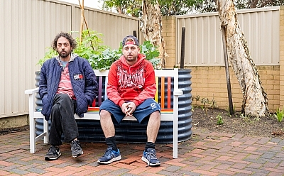 Two men sit on a colourful outdoor bench in front of a garden patch.
